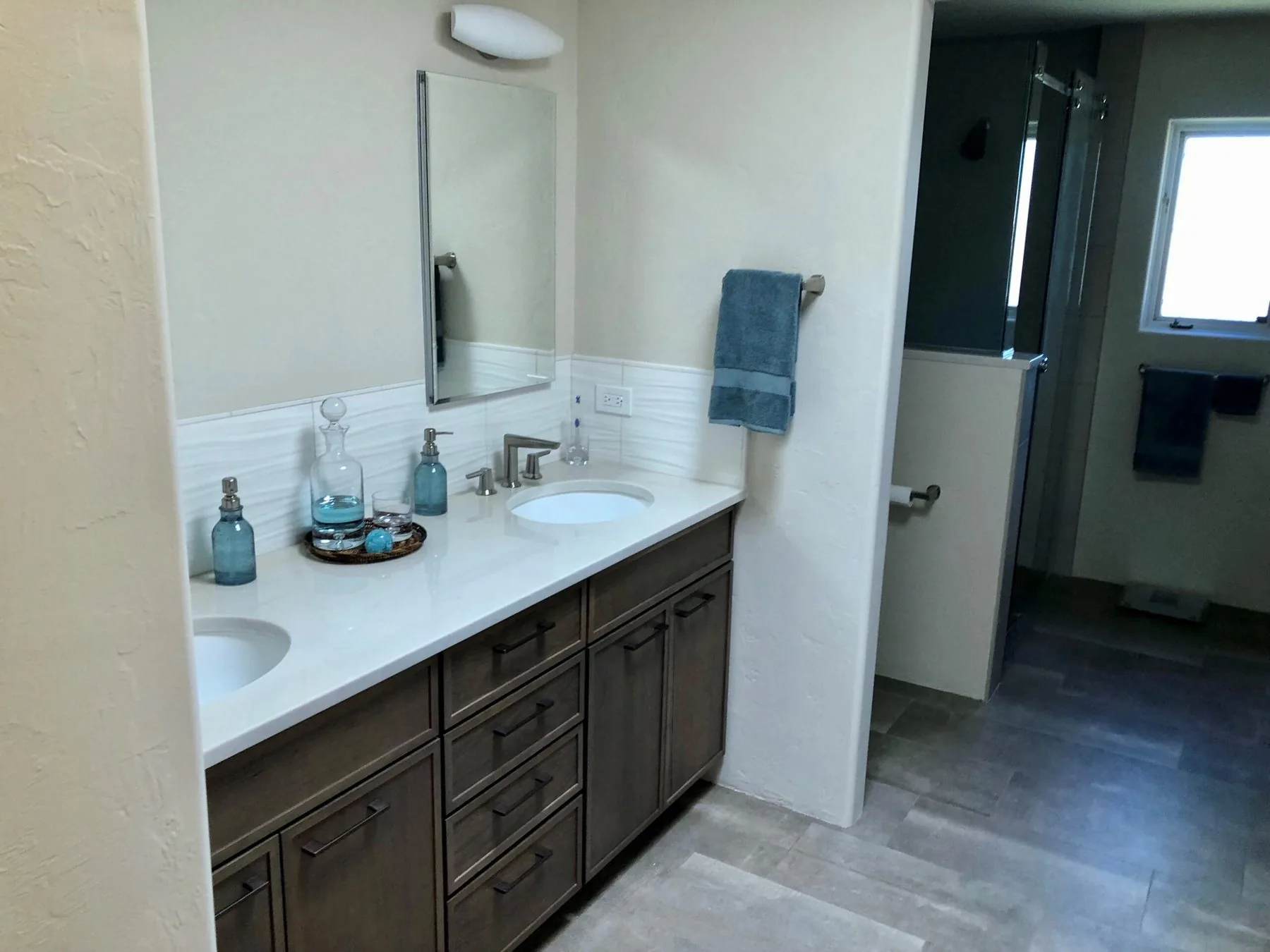 A remodeled bathroom with a double vanity, quartz counters, textured tile backsplash and a tiled walk-in shower